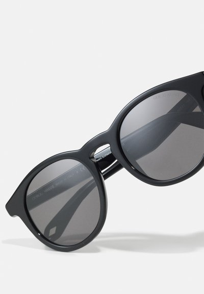 Occhiali da sole neri con montatura curva, lenti solide e finitura opaca. Presentano un branding discreto e un design unico con ritagli sulle stanghette.