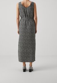 Femme portant une robe midi sans manches noire avec un petit imprimé floral blanc et des chaussures à talons blocs grises, se tenant devant un fond uni.