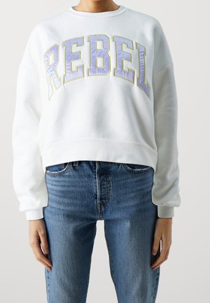 Personne portant un sweat-shirt blanc avec « REBEL » en grandes lettres lavande et un jean bleu, debout, les bras relâchés le long du corps.