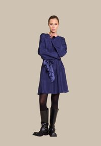 Mørkeblå satin kjole med lange ermer og knyttet liv, kombinert med knelange svarte lærstøvler med spenner.