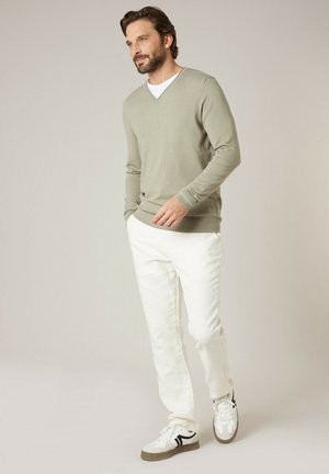 Homme barbu portant un pull à col en V beige, un T-shirt blanc, un pantalon blanc et des baskets blanches avec des détails noirs, debout devant un fond uni.