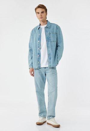 Chaqueta de mezclilla azul clara con bolsillo frontal, combinada con jeans azul claros a juego y una camiseta blanca. Los zapatos son cremosos con suelas marrones.
