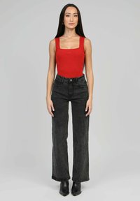 Femme à cheveux longs et sombres portant un haut rouge sans manches, un jean large taille haute noir et des bottes noires à bout pointu, se tenant devant un fond neutre.