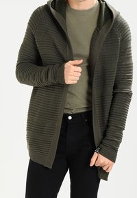 Olivgrön, texturerad stickad cardigan med huva, öppen front och förlängda ärmar. Bärs över en åtsittande mörk skjorta och svarta byxor.