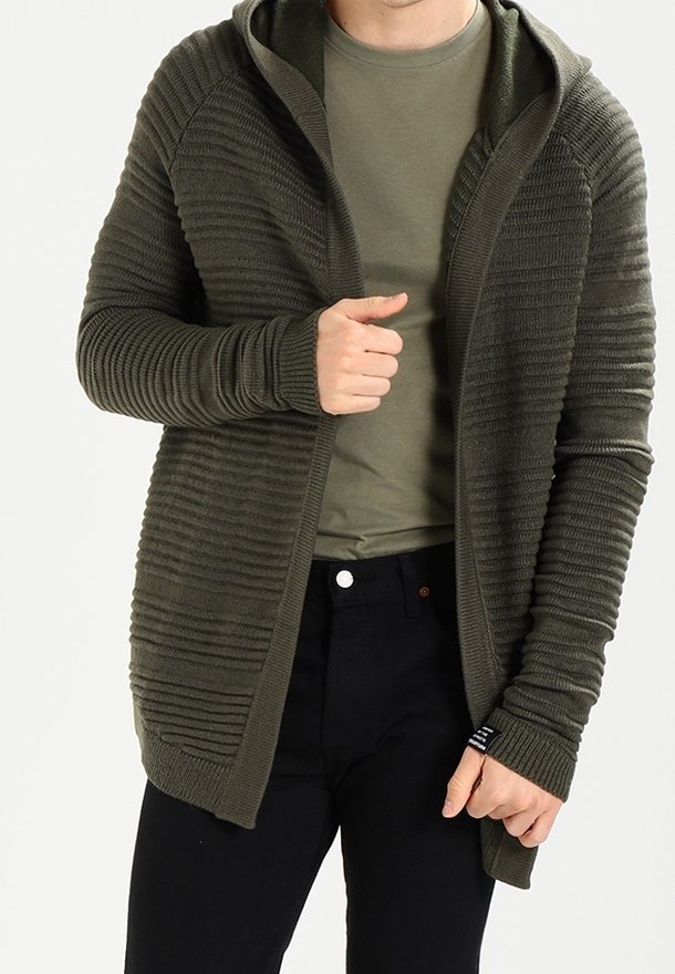 Olivgrön, texturerad stickad cardigan med huva, öppen front och förlängda ärmar. Bärs över en åtsittande mörk skjorta och svarta byxor.