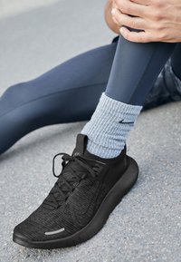 Černá sportovní bota s strukturovaným svrškem, plochými šňůrkami a reflexními detaily. Nosit s světle modrými žebrovanými ponožkami a šedými legínami, na šedém povrchu.