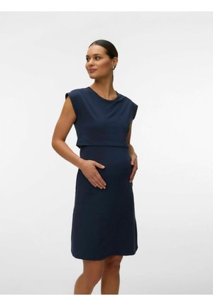 Gravid kvinde i en ærmeløs mørkeblå kjole står med hænderne på maven og kigger til siden mod en ensfarvet hvid baggrund.