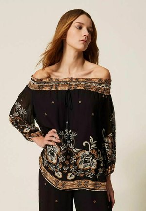 Donna con capelli castani lunghi che indossa una camicetta nera scollata con motivi floreali beige e bianchi, in piedi con una mano sui fianchi.