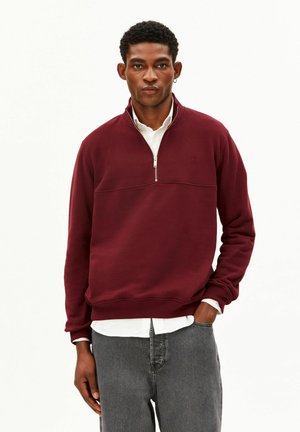 Junger Mann trägt einen burgunderfarbenen Halb-zip-Sweatshirt über einem weißen Hemd und grauen Jeans und steht mit einer Hand in der Tasche.