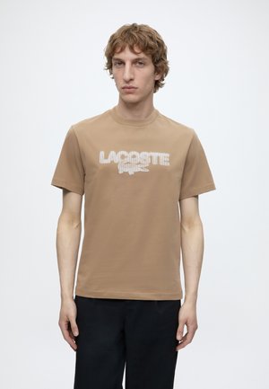 Jonge man met lichtbruin krullend haar, die een beige Lacoste t-shirt en zwarte broek draagt tegen een effen lichte achtergrond.
