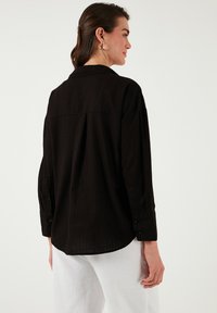 Black, long-sleeve button-up shirt featuring a relaxed fit, back yoke detail, and textured fabric. Buttons on sleeves, curved hemline.