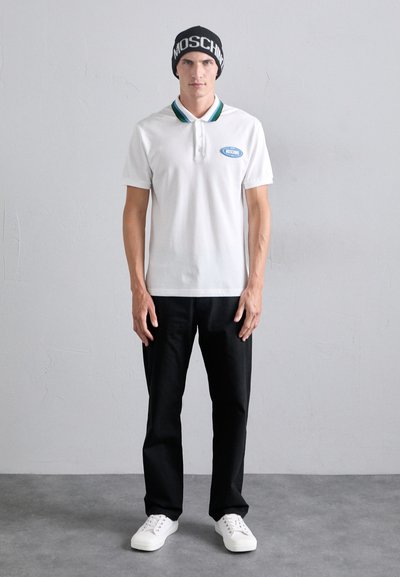 Valge polo särk roheliste ja siniste kaeluse aksentidega, mustad püksid ja valged tennised. Must kudumüts, millel on "MOSCHINO" tekst.