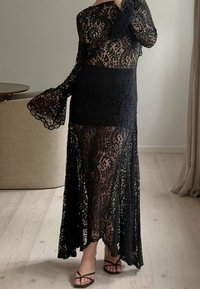 Juoda nėriniuota suknelė su ilgomis varpų formos rankovėmis ir prigludusia liemene, turinti sudėtingus gėlių raštus ir permatomą ilgą sijoną.