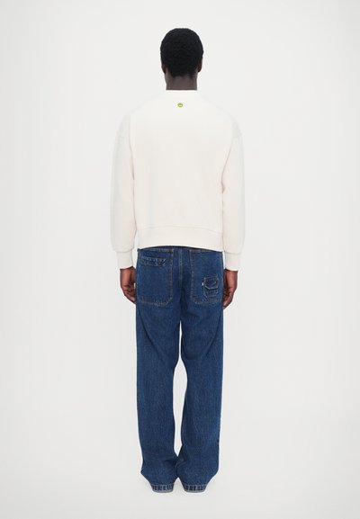 Sweatshirt blanc avec un col rond et des poignets côtelés, associé à un jean en denim bleu à coupe ample avec des poches plaquées et des éléments usés.