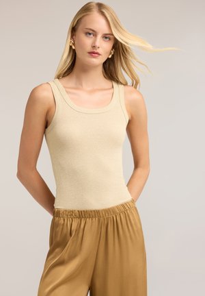 Donna bionda con orecchini a cerchio dorati, indossa un top beige senza maniche e pantaloni marroni larghi su uno sfondo neutro.