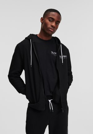Hanorac negru cu fermoar, cu șnururi albe, buzunare frontale și logo pe piept, purtat peste un tricou negru. Material din bumbac, croială lejeră.