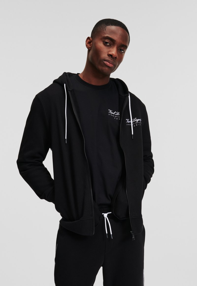 Zwarte zip-up hoodie met witte trekkoorden, voorzakken en logo op de borst, gelaagd over een zwart t-shirt. Katoenen materiaal, casual pasvorm.