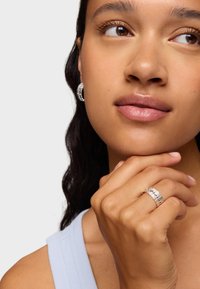 Anillo de plata con diseño en capas y ondulado; llevado en una mano de piel clara. El modelo tiene el pelo oscuro y ondulado y lleva pendientes de aro.