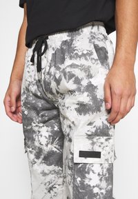 Person in Schwarz-Weiß-Tie-Dye-Cargohosen mit Seitentasche und schwarzem Kordelzug, kombiniert mit einem schwarzen Shirt, Hände entspannt an den Seiten.