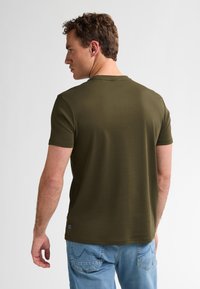 Olivgrünes T-Shirt mit kurzen Ärmeln und rundem Halsausschnitt. Der Stoff wirkt glatt und enganliegend, mit einem kleinen Markenetikett auf der linken Seite.