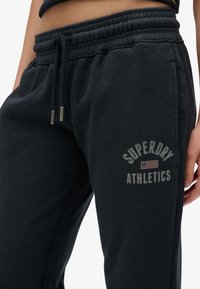 Close-up van zwarte sweatpants met een trekkoord in de taille en de tekst "Superdry Athletics" en een klein vlaglogo op de bovenbenen.