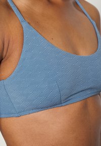 Bralette azul com padrão de espinha de peixe texturizado, alças finas e decote curvo. O tecido parece elástico e suave.