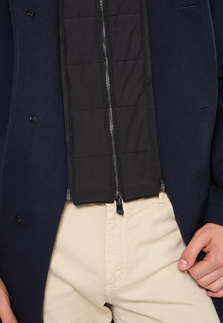 Manteau en laine marine avec un intérieur en nylon noir matelassé. Doté d'une fermeture éclair à l'avant et de boutons-pression. Associé à un pantalon en velours côtelé beige.
