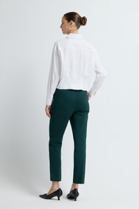 Camicia cropped bianca con collo, maniche lunghe, abbinata a pantaloni aderenti verde scuro e scarpe nere con tacco. Design semplice e sartoriale.