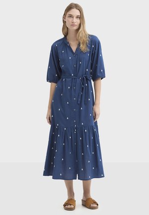 Vestido azul con botones, mangas cortas, cintura ceñida, falda de volantes y bordados blancos. Combinado con sandalias marrones.