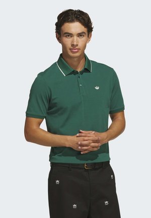 Млад мъж с тъмна коса, облечен в зелена поло тениска и черни панталони с бели бродирани лога, стои със събрани ръце отпред.