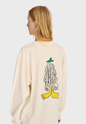 Giovane con lunghi capelli biondi indossa una maglietta crema con una figura dei cartoni animati con piedi gialli e cappello verde sulla schiena.