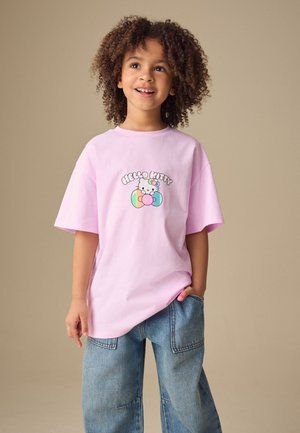 Lächelndes Kind mit lockigem Haar, das ein hellrosa Hello Kitty T-Shirt und blaue Jeans trägt, steht mit einer Hand in der Tasche.