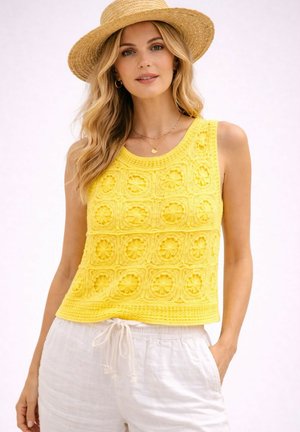 Femme portant un haut jaune au crochet sans manches, un pantalon blanc à cordon, un chapeau de paille et des bijoux en or, debout devant un fond uni.