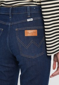 Dunkle Jeans aus Denim mit einem Lederlogo von Wrangler und gelben Nähten. Verfügt über ein Zickzackdesign auf den Gesäßtaschen und ein gestreiftes Oberteil.