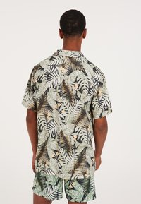 Camicia a maniche corte con motivo a foglie tropicali in verde, nero e beige. Tessuto leggero, vestibilità ampia, con colletto e chiusura a bottoni.