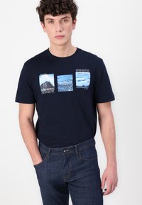 Donkerblauwe t-shirt met korte mouwen met drie rechthoekige grafische prints met natuurfoto's, gelabeld met "TOM TAILOR" en "RESORT." Denim jeans.