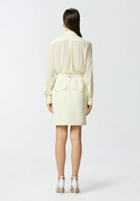Blusa gialla trasparente a maniche lunghe con vita aderente, abbinata a una gonna gialla chiara a lunghezza ginocchio con tasche posteriori e cordoncino.