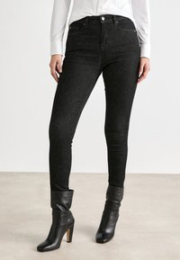 Černé skinny džíny vyrobené z pružného denimu, s vysokým pasem a klasickým designem pěti kapes. V kombinaci s upnutými černými kotníkovými botami.