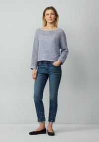 Hellblauer, gestrickter Pullover mit weitem Ausschnitt und langen Ärmeln. Das Model trägt dunkle Jeans mit Perlendetails am ausgefransten Saum und schwarze Ballerinas.