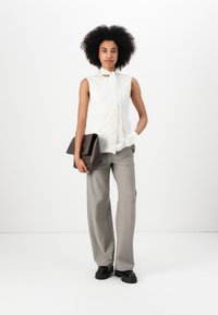 Weißes ärmelloses Bluse mit einem Bindedetail, kombiniert mit hellgrauen weiten Hosen. Das Outfit wird durch eine dunkelbraune Clutch-Tasche ergänzt.