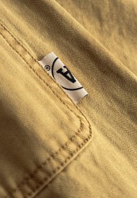 Beiges Stoff mit Fischgrätmuster, versehen mit einer abgesteppten Tasche und einem Logetikett mit einem kreisförmigen Rand und dem Buchstaben "A".