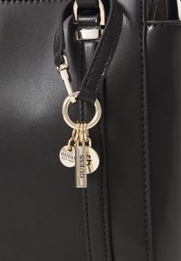Sac à main en simili cuir noir avec une finition brillante, ferrures dorées et plusieurs accents de porte-clés avec des logos de marque.