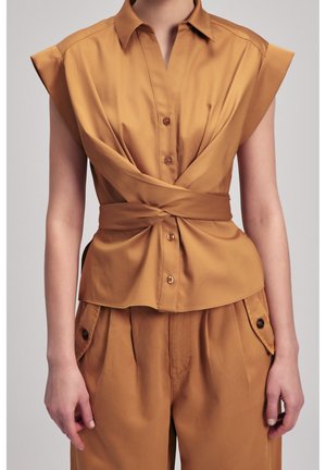 Beżowa bezrękawnikowa bluzka na guziki z szerokim kołnierzem i paskiem przewiązywanym wokół talii, zestawiona z pasującymi beżowymi spodniami z wysokim stanem i kieszeniami na klapki.