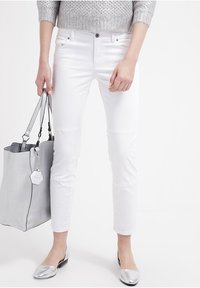 Pantalones ajustados blancos con una textura suave, combinados con un bolso de cubo gris claro y bailarinas plateadas brillantes. El bolso tiene un acabado texturizado y una etiqueta en forma de hexágono.