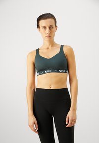 Sujetador deportivo verde oscuro con un diseño de escote en V, tirantes anchos y una banda negra con el logo de Nike. Combinado con leggings negros de cintura alta.