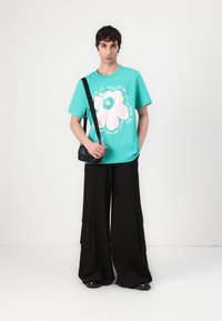 Turquoise cotton t-shirt with a large white flower graphic, paired with black wide-leg trousers featuring cargo pockets and a small black bag.