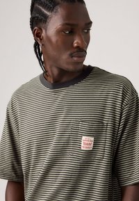 Ριγέ κοντομάνικη μπλούζα σε χακί και μαύρο, με θήκη στο στήθος που φέρει κόκκινη και λευκή υφασμάτινη ετικέτα. Απαλό βαμβακερό υλικό.