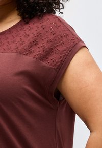 Κοντινό πλάνο ενός μπορντό κοντομάνικου τοπ με κεντητό δαντελένιο σχέδιο λουλουδιών στον ώμο, που φορά ένα άτομο με σγουρά μαλλιά.