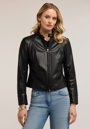 Lyshåret kvinde iført en sort læderjakke, lyseblå jeans, guldringede øreringe og armbånd, stående mod en ensfarvet baggrund.