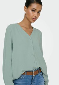 Femme portant des boucles d'oreilles créoles argentées, un chemisier vert clair fluide à col en V boutonné, un jean bleu et une ceinture marron sur fond blanc.